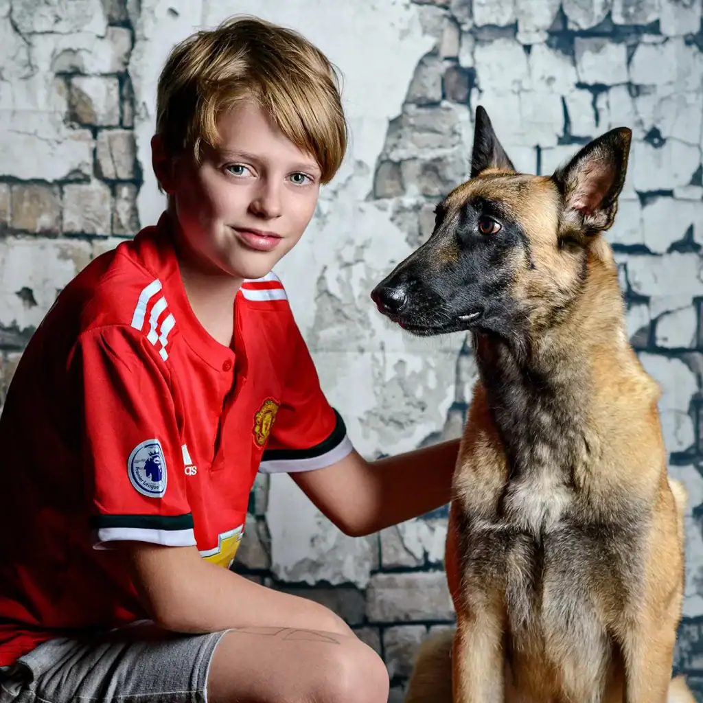 Young boy in red football shirt posing with Belgian Malinois dog against a textured brick wall backdrop