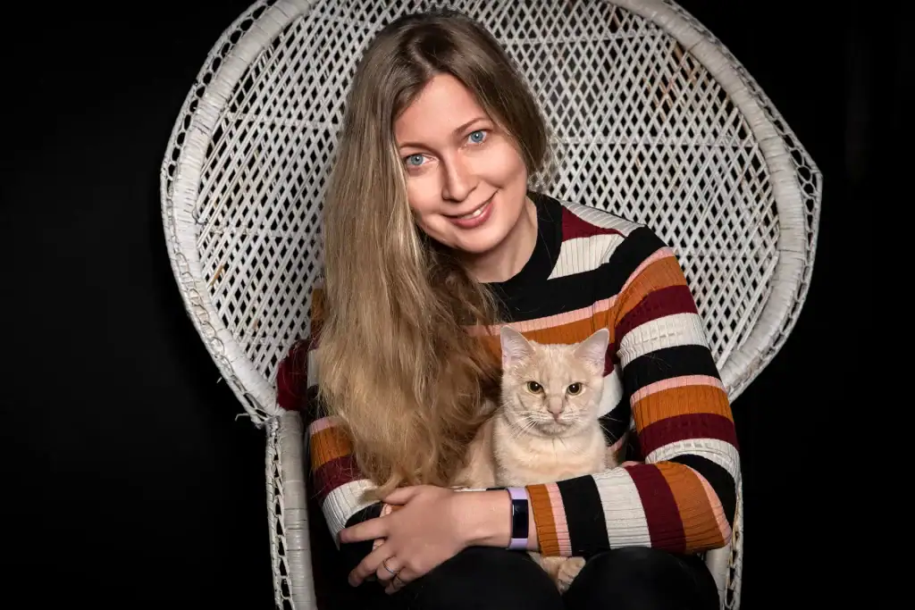 Smiling woman with long blonde hair holding her cream-coloured cat in a white wicker chair, captured in a professional pet studio shoot