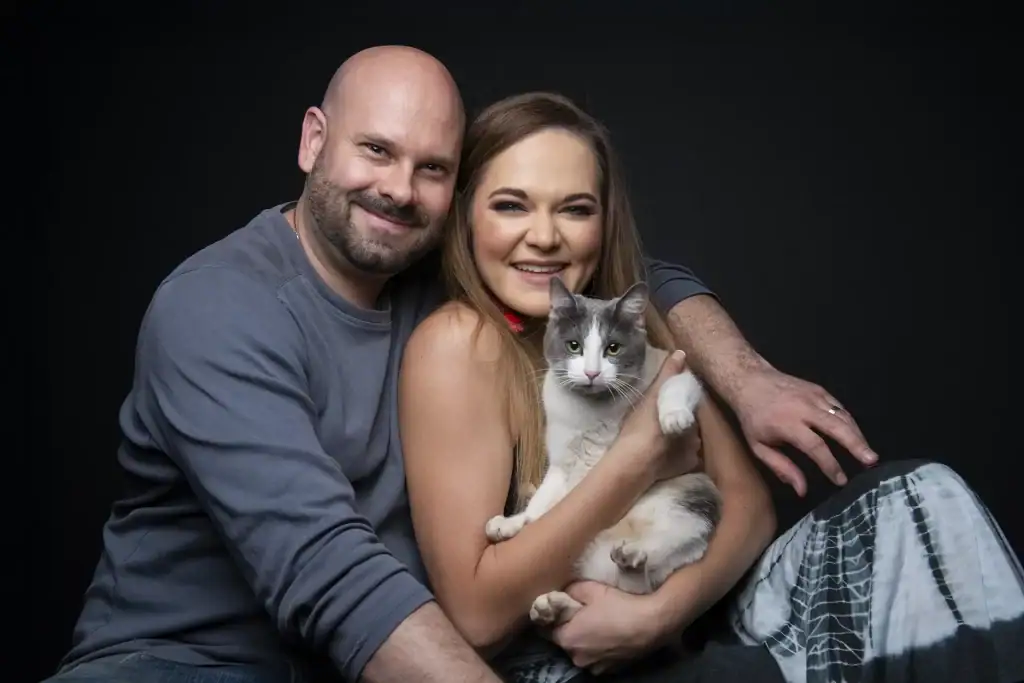 Smiling couple embracing a white and grey cat in a studio portrait with a black background