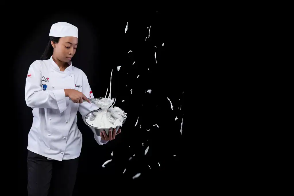 Black female culinary student in full chef uniform whisking batter mid-motion, photographed in studio with black background for branding and corporate events