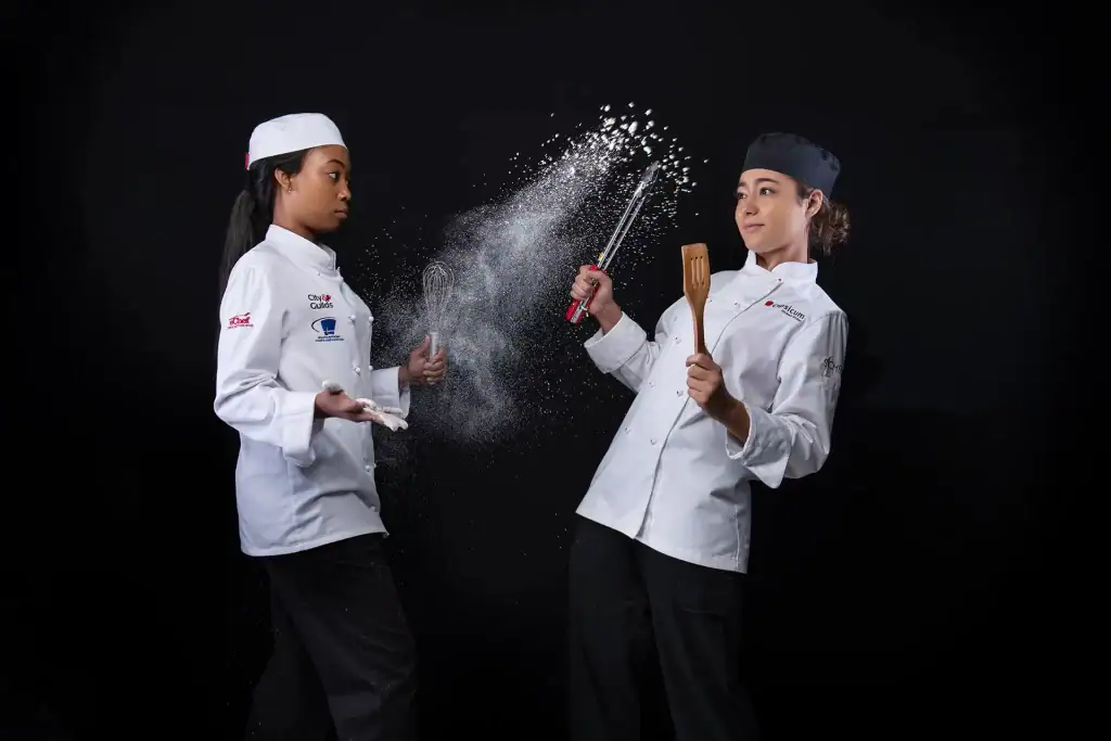 Two female culinary students playfully throwing flour during a chef school photoshoot, dressed in professional white chef uniforms with black background