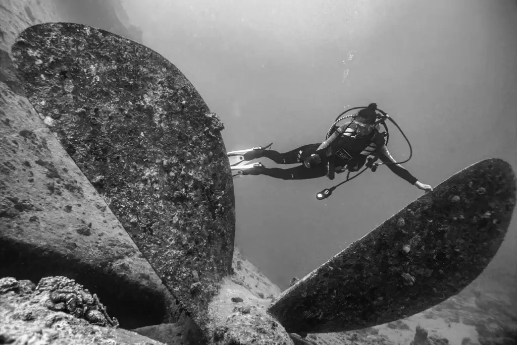 Black and white photo of a scuba diver examining a giant propeller on a sunken shipwreck during an underwater exploration dive