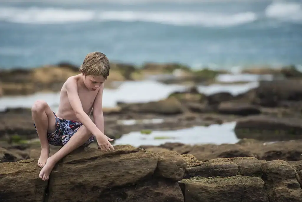 Young boy in swim shorts exploring rocks and tidal pools on a rugged coastline, captured for an editorial nature or travel story