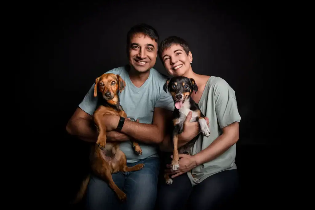 Happy couple posing in a photography studio with two small dogs, all wearing soft blue and green tones against a black background