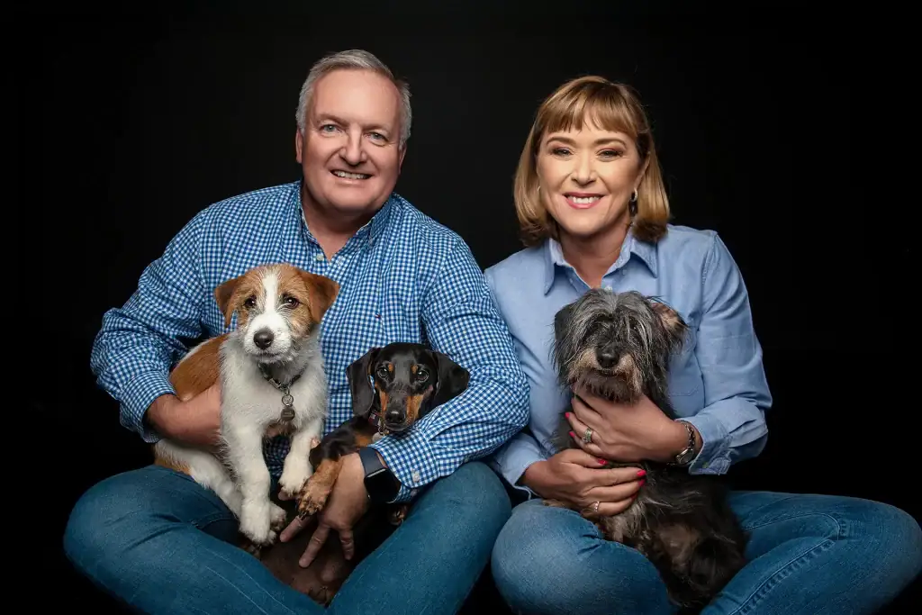 Smiling couple in studio holding three dogs of different breeds, all looking at the camera against a black background