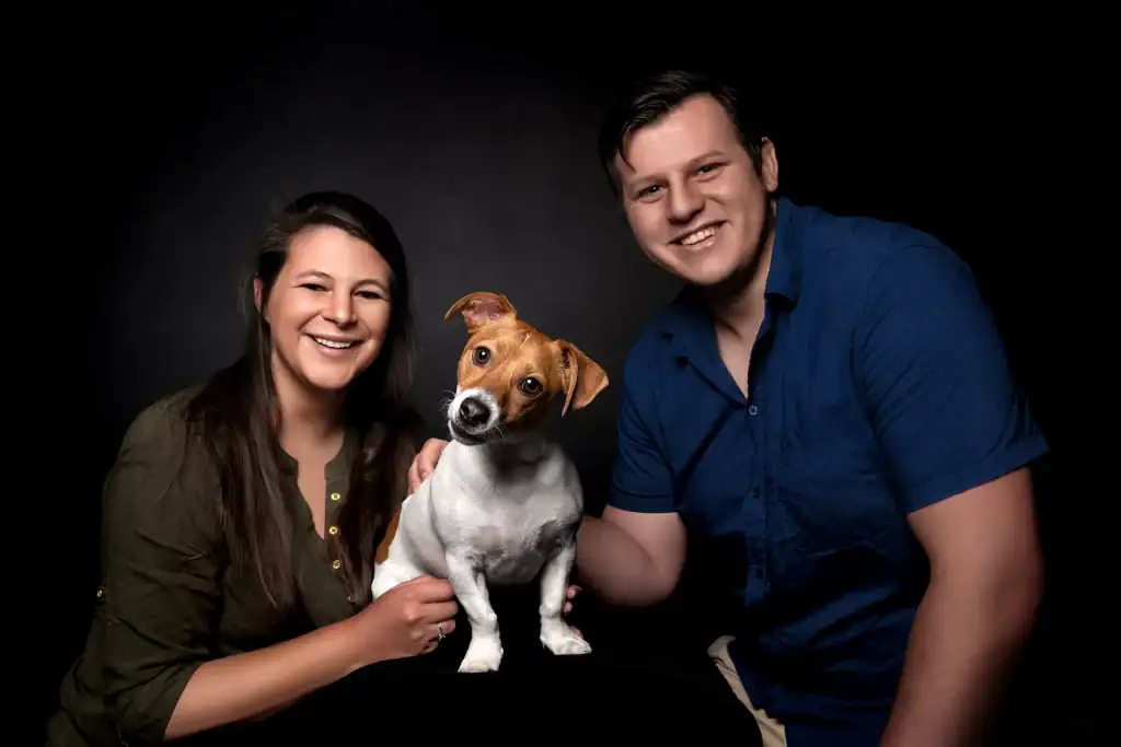 Happy couple posing in a dark photo studio with their Jack Russell Terrier, all smiling towards the camera