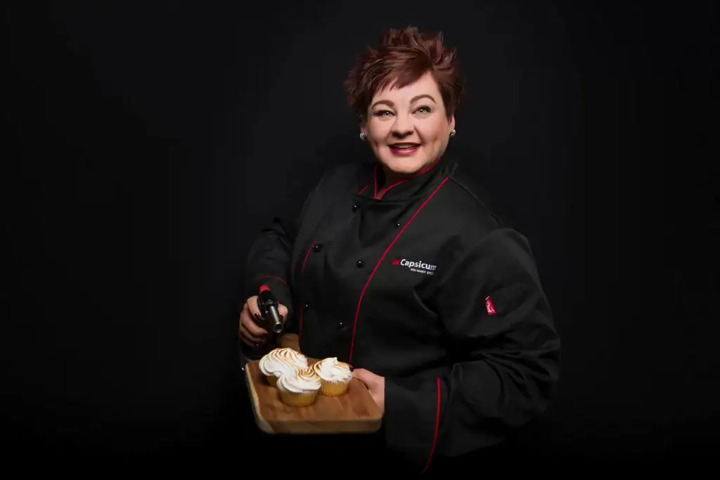 Female chef in black and red Capsicum Culinary Studio uniform smiling while toasting meringue cupcakes with blowtorch on black background