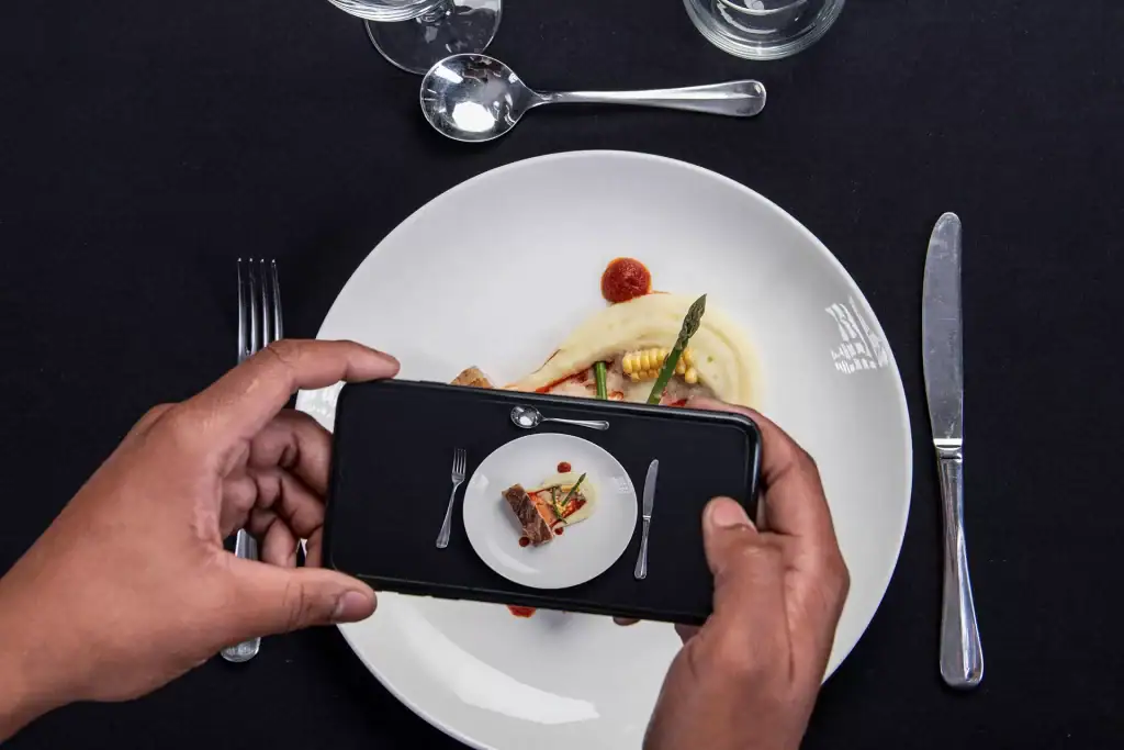 Culinary student capturing gourmet dish with smartphone creating tiny plate illusion in professional food photography setup