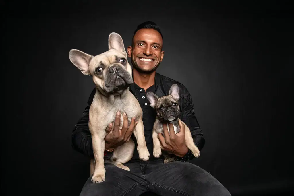 Smiling man holding two French bulldogs, one adult and one puppy, in a professional studio portrait against a dark background