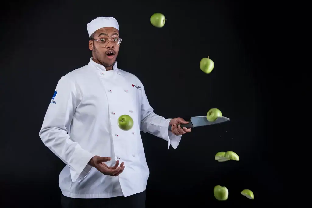 Black male culinary student juggling green apples with a knife during a fun chef photoshoot, wearing white uniform and hat on a black background