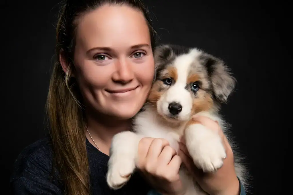 Young woman smiling warmly while holding a fluffy Australian Shepherd puppy with blue eyes against a black studio background