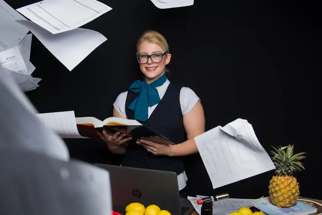Blonde hospitality student in uniform holding a textbook while papers fly through the air, styled corporate portrait with creative flair on black background