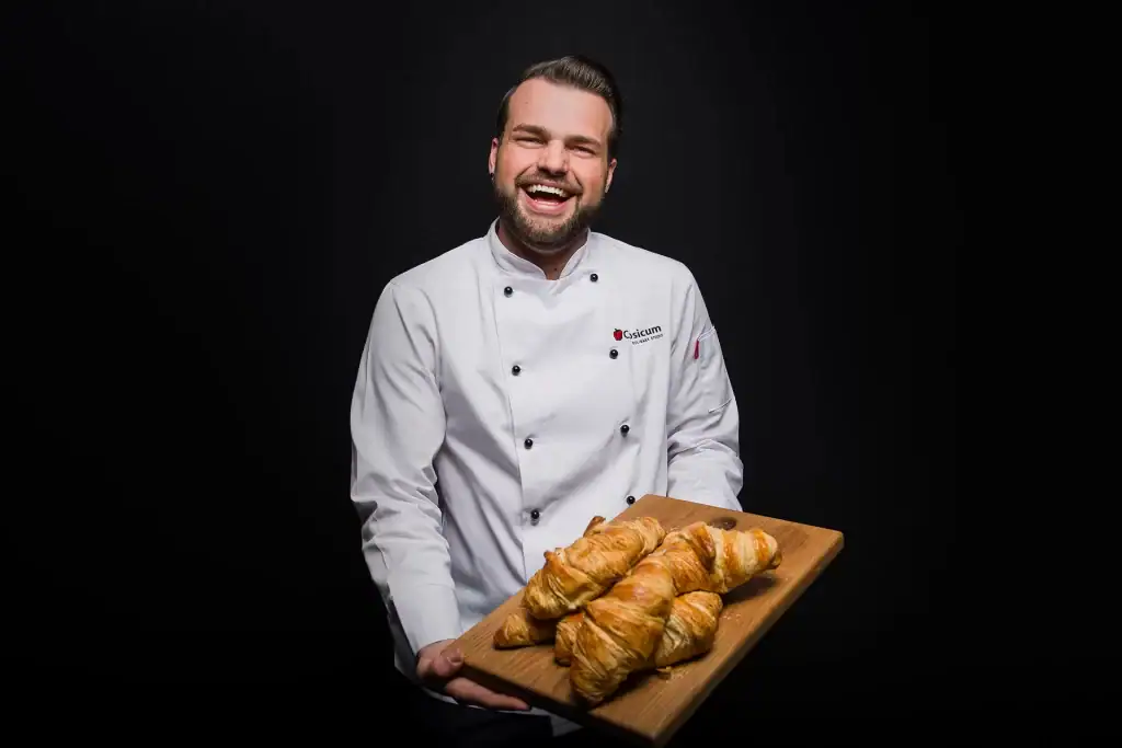 Smiling male chef in white Capsicum Culinary Studio uniform holding a wooden board of golden croissants on black background