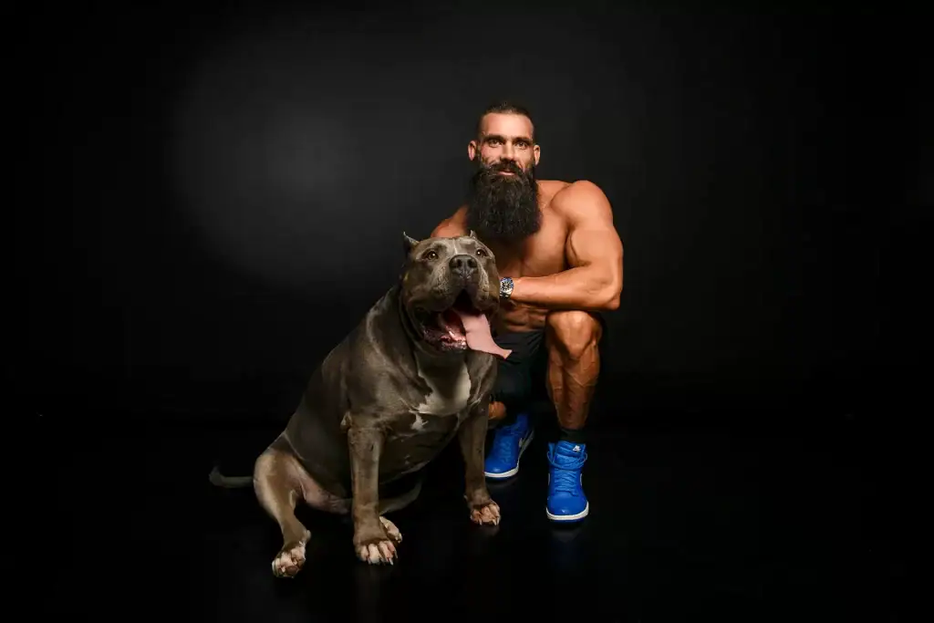 Muscular man with full beard posing shirtless beside his large American Bully dog in a dramatic black studio setting