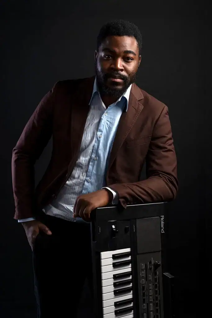 Professional headshot of a man in a blazer leaning on a Roland keyboard, photographed in a studio with dark background, ideal for corporate, personal branding, or music promotion