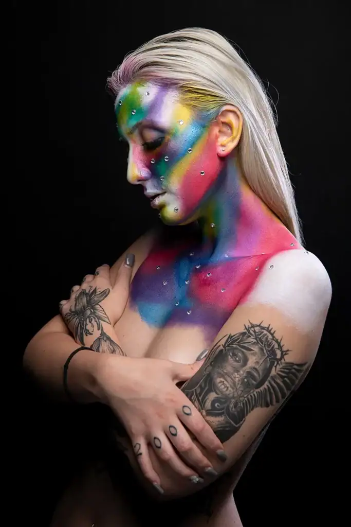 Artistic nude portrait of a woman with vibrant rainbow body paint, rhinestone embellishments, and tattoos on arms, shot in professional studio lighting