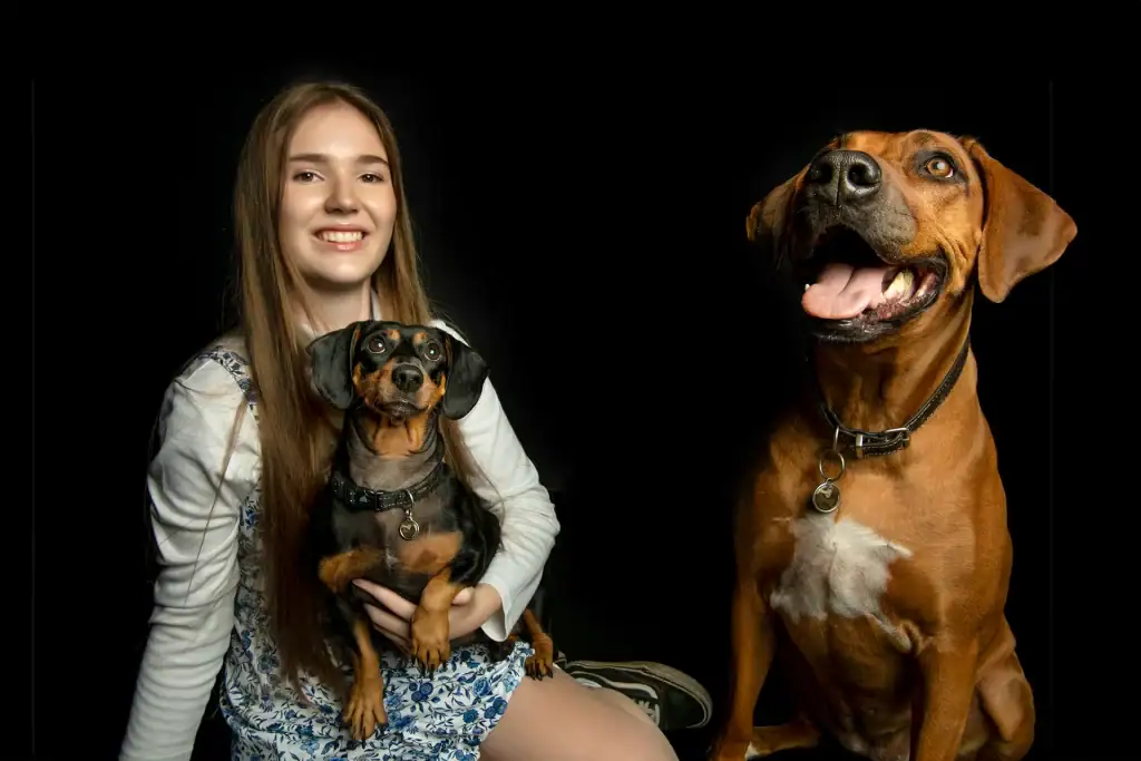Smiling girl holding a dachshund beside a happy ridgeback, captured in a professional studio pet portrait session with a black background