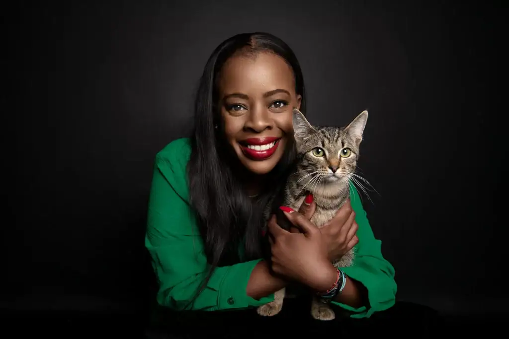 Studio portrait of a smiling woman in a green top holding her tabby cat against a black background, professional pet and owner photography