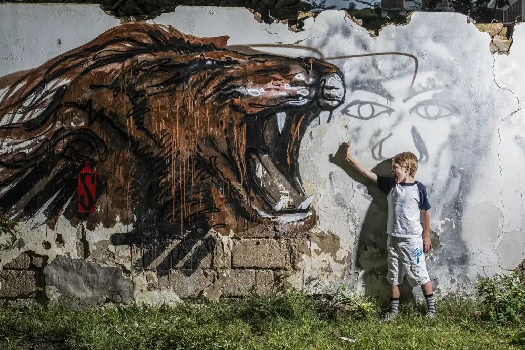 Young boy posing against a crumbling wall with vivid street art of a roaring lion, ideal for urban portraiture and editorial lifestyle photography