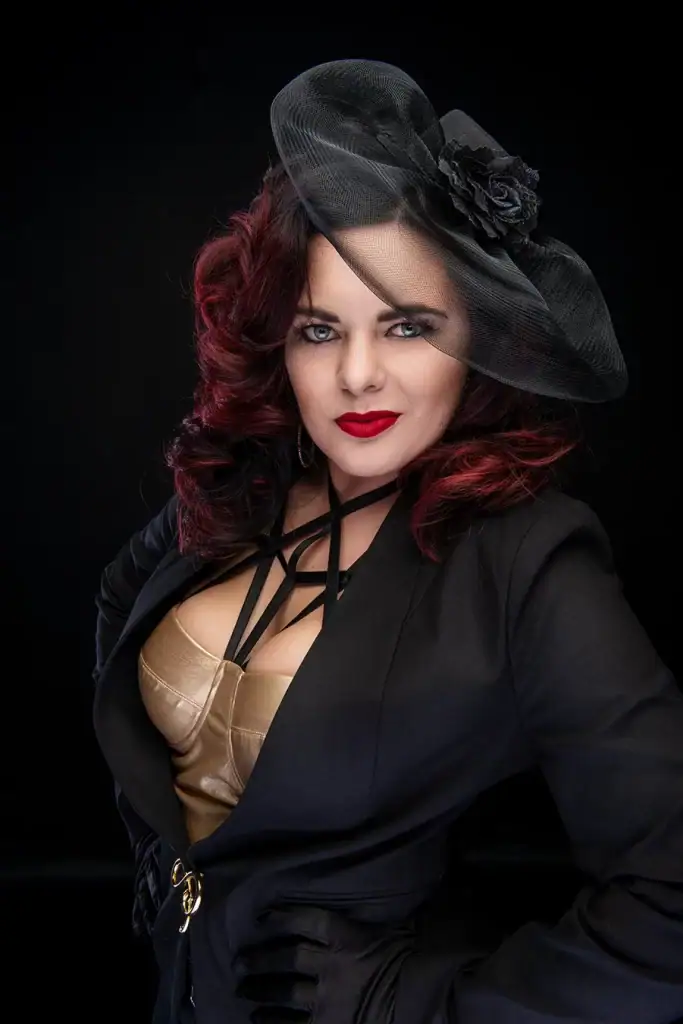 Stylish woman with red curls wearing a black fascinator, gold corset, and tailored blazer poses confidently in a studio portrait with bold makeup and elegant flair