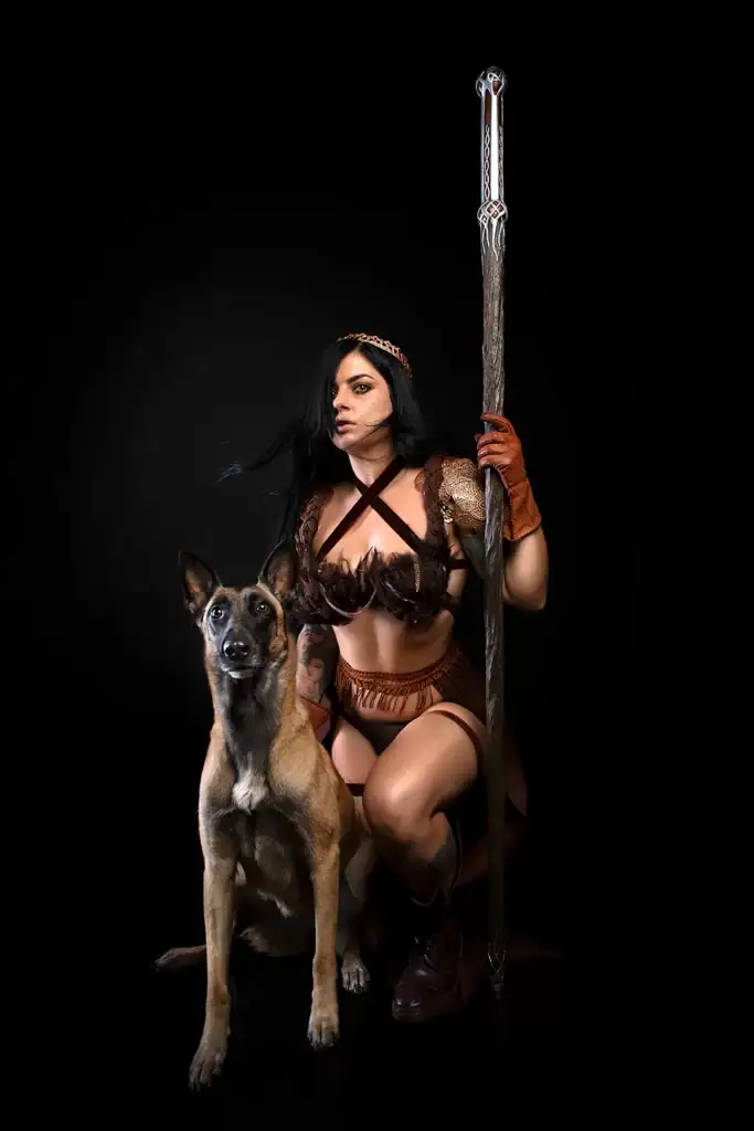 Fierce female warrior in fantasy cosplay kneeling beside Belgian Malinois dog holding staff in dramatic dark studio portrait