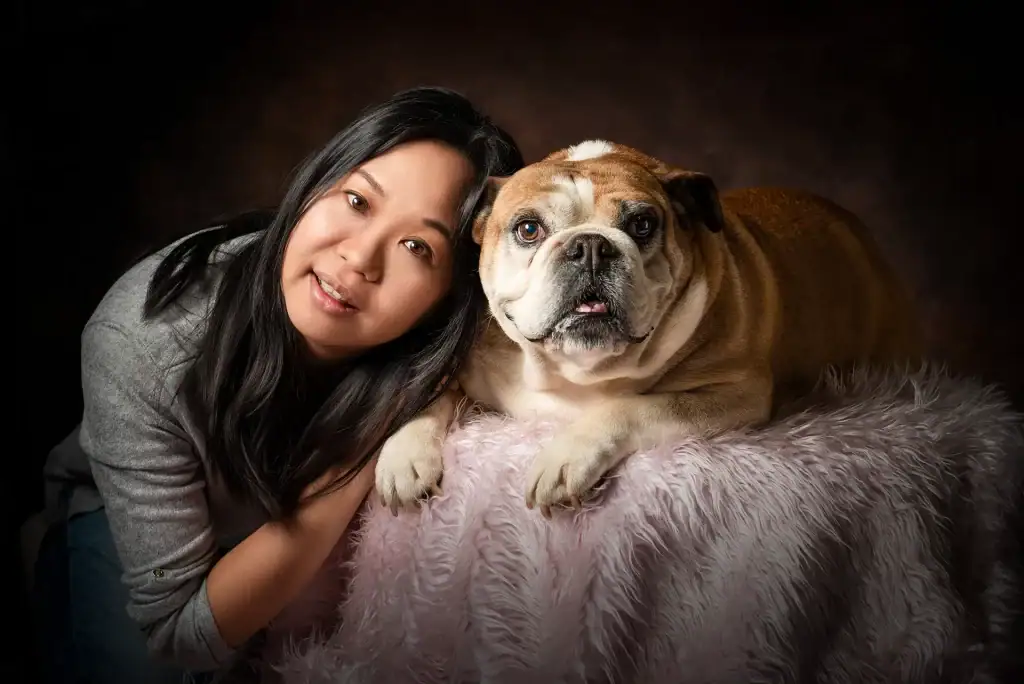 Woman with long dark hair leaning close to her English bulldog on a pink fluffy blanket in a studio portrait with brown backdrop