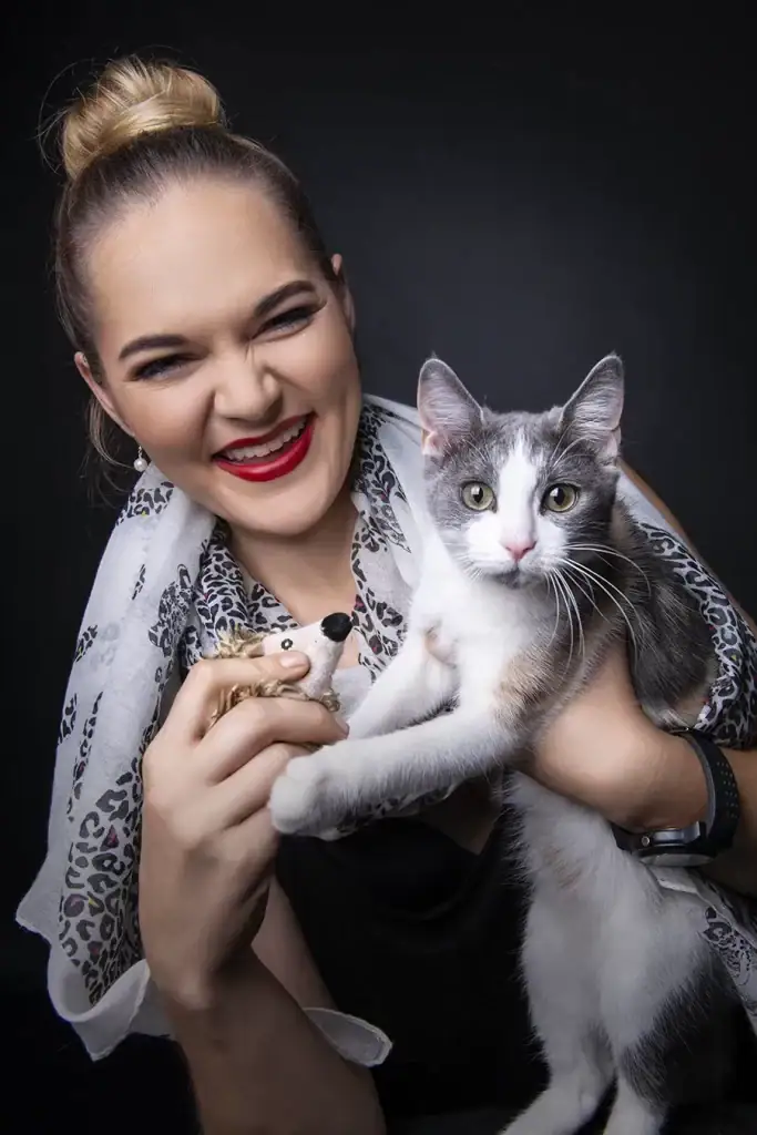 Playful studio portrait of a woman laughing while holding a grey and white cat and a small toy hedgehogv