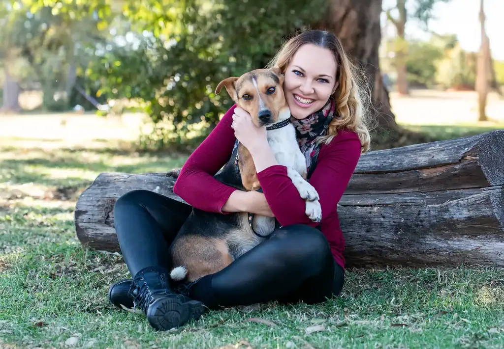 Smiling woman in burgundy top sitting on the grass, hugging her dog in a sunlit park with trees and a log in the background