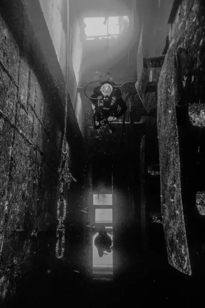Black and white underwater photograph of a scuba diver swimming through the interior of a sunken shipwreck, framed by metal walls and soft light