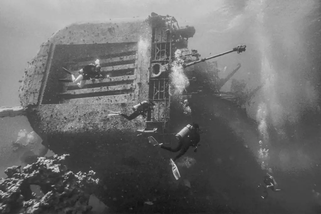Black and white underwater photograph of scuba divers exploring a sunken warship wreck with visible gun mounts and large hull structure