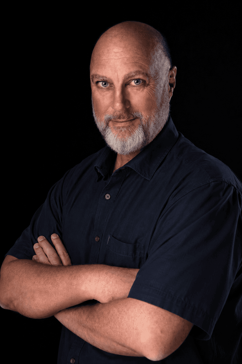 peter-driessel-professional-photographer-johannesburg Portrait of a bald, bearded man with blue eyes, wearing a navy button-down shirt with arms crossed against a black background.